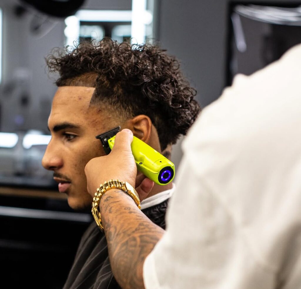 Interior view Cut Throat Barbershop featuring a professional barber in a white graphic t-shirt preparing a station with a classic black leather barber chair and large illuminated mirror.